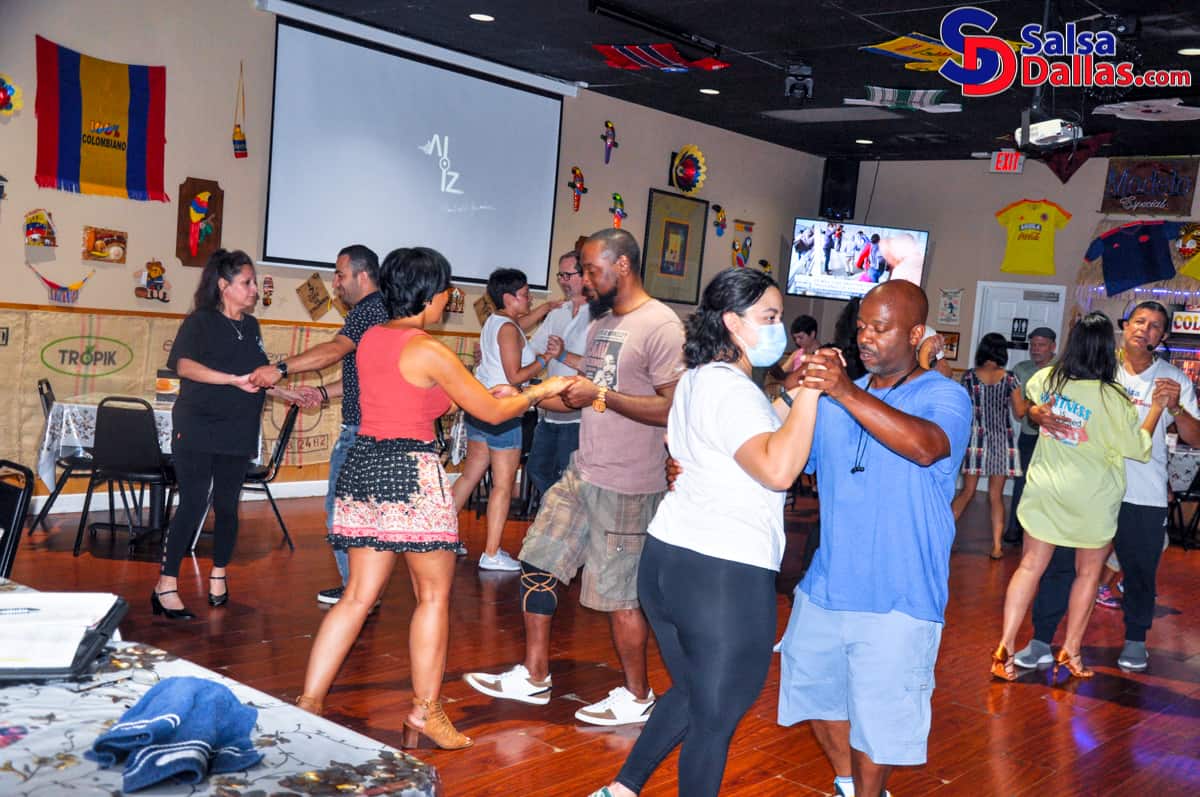 Vibrant social dance scene at Salsa Dallas with couples practicing salsa and bachata moves in a lively, colorful nightclub decorated with Colombian flags and salsa-themed artwork.