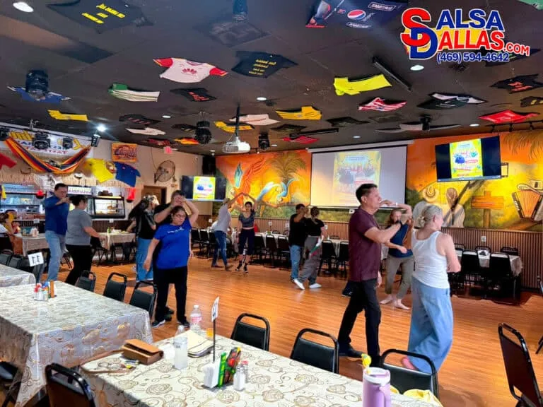 Vibrant dance class at Salsa Dallas restaurant with participants practicing salsa moves on a wooden floor, colorful mural walls, and lively decor including international flags on the ceiling.