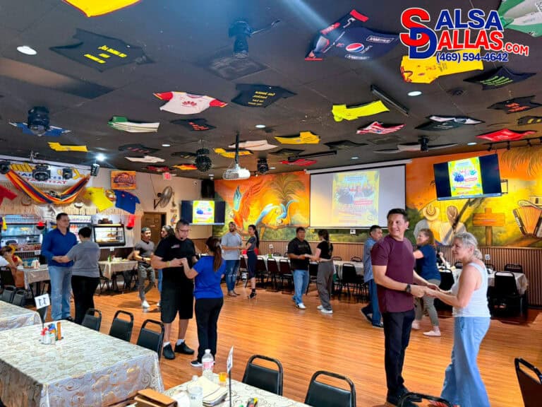 Dance class at Salsa Dallas with couples practicing salsa moves in a lively, colorful studio with tropical murals and decorated ceiling featuring sports jerseys.