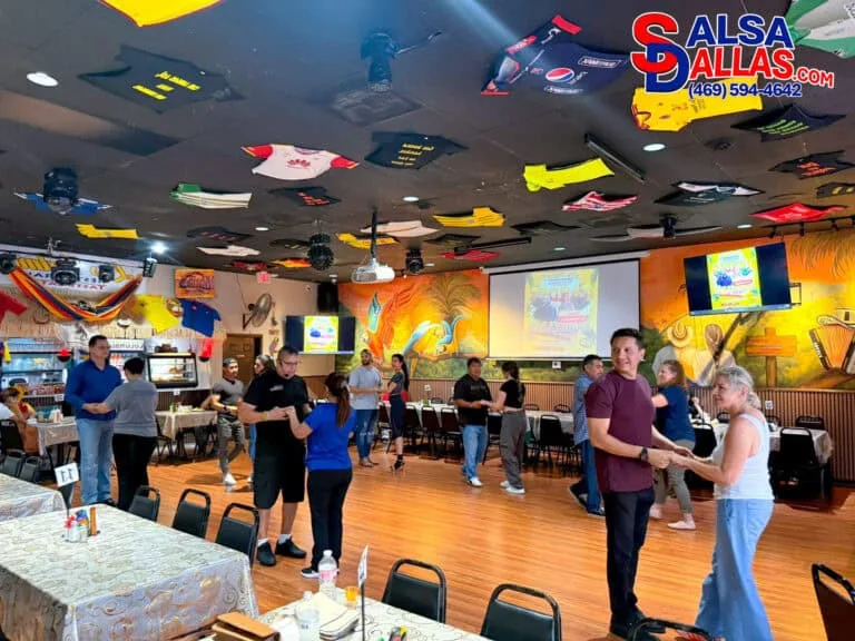 Dance class at Salsa Dallas with couples practicing salsa moves in a lively, colorful studio with tropical murals and decorated ceiling featuring sports jerseys.